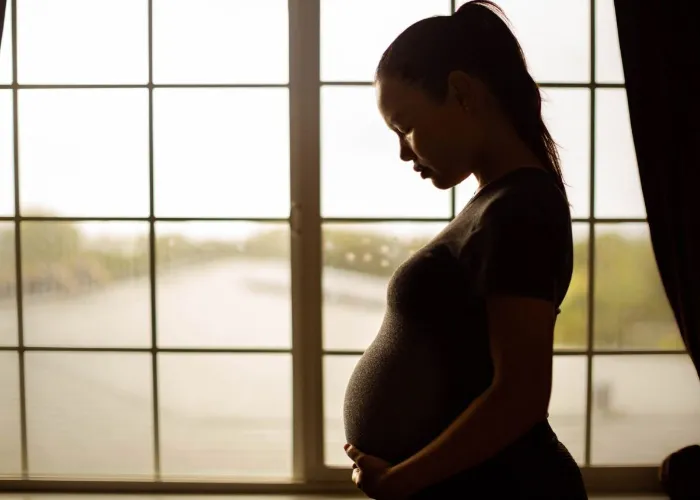 A sad pregnant woman standing near a window looking down depressed.