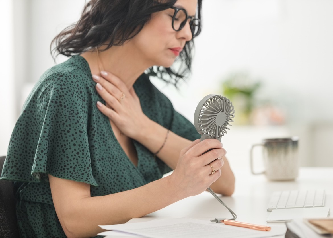 Menopausal mature woman wearing green dress sitting at the desk in the office, having hot flashes and using hand fan.