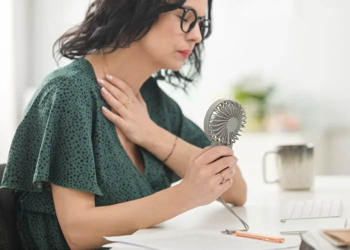 Menopausal mature woman wearing green dress sitting at the desk in the office, having hot flashes and using hand fan.