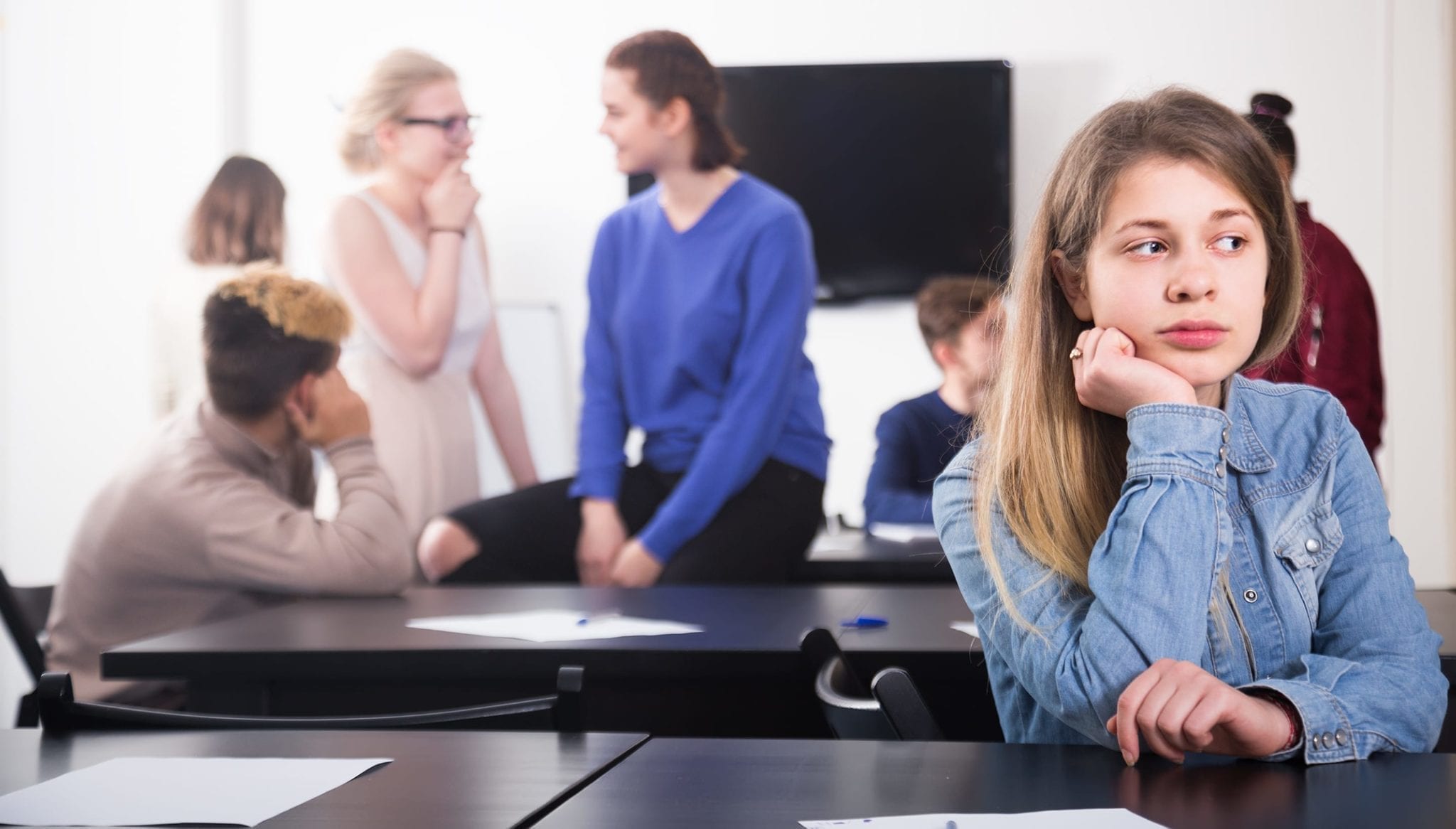 Image of a teenage girl sitting at a desk by herself while all of her classmates hang out in the background.