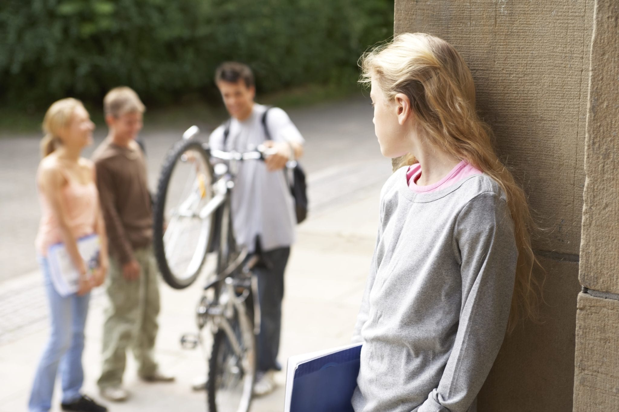Image of teenage girl leaning against a wall while looking over at three of her classmates hanging out without her.