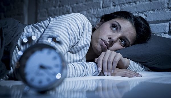 Image of a woman lying in her bed wide awake with an alarm clock in the front of the camera.