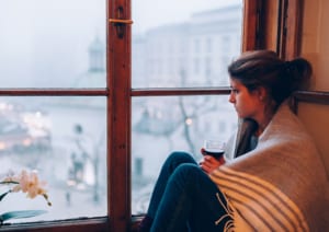 Image of a woman with a blanket wrapped around her sitting next to a window, with a glass of wine.