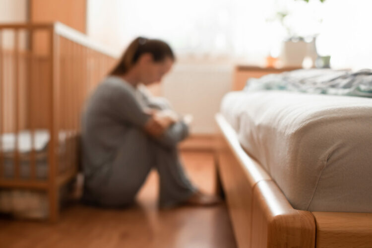A mother sits on the floor by a crib showcasing postpartum symptoms not to ignore.